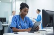 © Raisa - African American nurse checks schedule on digital tablet in medical office. Healthcare specialist works with tech at hospital desk. Woman wears stethoscope, uniform. Doctors on background, clinical