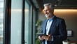 © Pete - Smiling middle-aged businessman in suit using digital tablet. Mature executive manager looking away. Happy ceo holding tech device in modern office. Professional corporate leader in successful job.