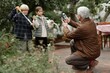 © Seventyfour - Senior Caucasian man kneeling outdoors photographing two children blowing soap bubbles in village garden, kids smiling and interacting with each other while man using smartphone