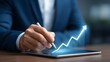 © sbgraphic - Businessman using a stylus on a tablet to interact with a glowing upward trending financial graph symbolizing growth and success