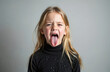 © Pete - Young girl showing disgust face expression with tongue out. Blonde hair female child portrait in studio, grey background. Nonverbal communication of hate, dislike, disapproval. Negative feeling