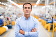 © Kowit - A confident man in a light blue shirt stands with crossed arms in a busy factory or industrial setting with blurred machinery in the background.