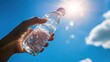 © Naknakhone - A hand holds a clear plastic water bottle against a bright blue sky with sunlight and scattered clouds.
