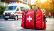© 24DIY - Red first aid bag with white cross emblem sits on the ground ambulance parked in background with flashing lights in
