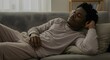 © Bhagya - Tranquil African American man in cozy pajamas resting on a comfortable living room sofa, eyes closed with a peaceful expression, enjoying a moment of serene relaxation.