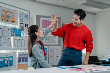 Fototapete - Two smiling Asian colleagues, a male and female UX/UI designer, high-fiving in their office while celebrating successful app design development in front of wireframes and prototypes