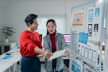 Fototapete - Two young Asian ui/ux developers reviewing mobile app design wireframes displayed on a whiteboard, discussing improvements and functionalities in their modern office