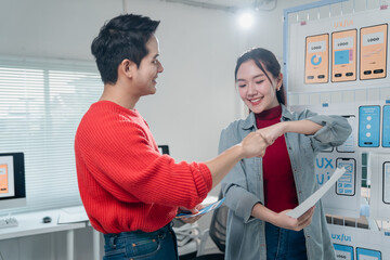 Fototapete - Two young asian ux/ui designers fist bumping and smiling after successfully completing a project, holding mockups and standing next to a whiteboard with ux/ui design sketches