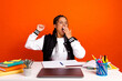 © deagreez - Young college student yawning while studying at desk with vibrant orange background and educational supplies