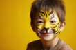 © firdan - A young child with tiger face paint smiles set against a vibrant yellow backdrop