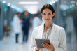 © antusher - Confident female doctor holding tablet standing in hospital hallway with colleagues and bright modern lighting