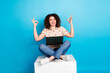 © deagreez - Young woman sitting on a cube with a laptop in her lap, gesturing okay and cheerful, set against a vivid blue background.