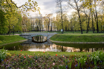 Poster - A serene park features a picturesque bridge crossing a tranquil river, bordered by colorful flowers and tall trees under a soft morning sky.