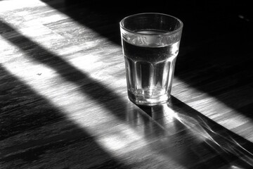 Poster - Close-up of a transparent glass of water placed on a wooden surface with dramatic shadows and light patterns creating a high-contrast monochrome scene