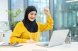 © Liubomir - A woman in a hijab celebrates success in an office setting, smiling at her laptop.
