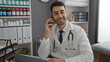 © Krakenimages.com - Hispanic male doctor in clinic office wearing white coat, smiling while talking on phone with medical files and shelves in blurred background suggesting professional healthcare setting.