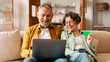 © Home-stock - Happy modern senior spouses shopping online via laptop with credit card, man and woman making internet purchases, sitting on couch
