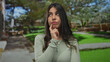 © Krakenimages.com - Young hispanic woman in green park outdoors, looking thoughtful with finger on chin and surrounded by lush scenery, suggesting a peaceful and reflective moment.