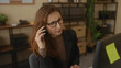 © Krakenimages.com - Young business woman in office talking on phone looking concerned with computer in front and shelves in background showing workplace interior setting.