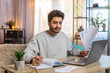 © Andrii Iemelianenko - Young Indian man freelancer sitting at table reviewing financial reports on laptop, holding documents and receipts, taking notes in notebook. Hispanic guy sits nearby at home, focused on his own task.