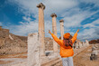 © EdNurg - Young woman tourist is posing with raised arms near ancient ruins of Asklepion, famous landmark in Bergama, Turkey