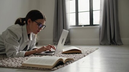 Fototapete - Diligent student wearing glasses is lying on a comfortable rug, typing on her laptop with an open textbook beside her, indicating focused learning or research in a cozy home environment
