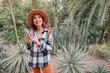 © EdNurg - Young woman using smartphone and enjoying exotic botanical garden, surrounded by cacti and succulents