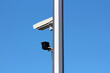 © hecos - Two surveillance cameras, one white and one black, are attached to a tall metallic pole, monitoring the area under a cloudless blue sky