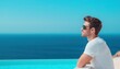 © Preeyanuch - A man in sunglasses and a white shirt gazes thoughtfully over a turquoise infinity pool towards a calm blue ocean under a clear sky.
