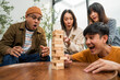 © Chanakon - Young Asian friends are happily playing wooden block together at home, enjoying their leisure time. Group friends carefully remove a wooden block while others watch, having fun a classic table game.