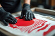 © Pete - Hands in black gloves apply red ink screen print of sun, waves design onto white fabric. This is close-up of custom apparel creation process, showcasing textile printing technique, craftsmanship.