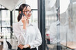 © kenchiro168 - Young Asian businesswoman with glasses standing indoors, smiling while talking on smartphone, wearing white shirt, modern office background, confident and professional mood
