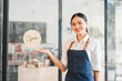 © kenchiro168 - Young Asian woman wearing denim apron smiling and standing in front of glass door with open sign, welcoming customers to modern cafe or coffee shop, friendly and inviting atmosphere