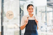 © kenchiro168 - Young Asian woman wearing denim apron standing in front of glass door with open sign, smiling confidently, welcoming customers to modern cafe or restaurant