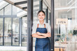 © kenchiro168 - Young Asian woman wearing denim apron standing at entrance of modern cafe, arms crossed, smiling confidently, welcoming customers, glass door with open sign, bright and inviting atmosphere