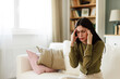 © Dorde - Young Woman in Green Sweater Sitting in Bright Living Room Holding Head in Pain