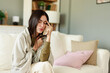 © Dorde - Young Woman Feeling Sick Sitting on Couch Wrapped in Blanket in Cozy Living Room