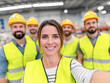 © Kowit - A smiling female construction worker takes a selfie with four male coworkers wearing yellow safety vests and helmets in a busy industrial setting.