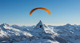 A vibrant orange paraglider soars gracefully above a breathtaking snow-covered mountain range under a clear blue sky.