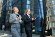© muse studio - Business professionals in smart attire engage with devices while walking in a modern urban setting during a sunny day
