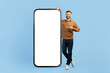 © Anastasiya - Smiling man pointing at big blank smartphone screen. Large mobile mockup ideal for digital ads, app showcase, or tech product presentations, male standing on blue studio background