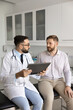 © fizkes - Serious Latin general practitioner man asking male patient about health complaints, listening, holding pad with medical records, history, giving consultation in clinic examination room
