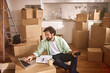 © Stockphotodirectors - A man sits comfortably in a new home surrounded by unpacked boxes, using a laptop to organize tasks and holding a phone in his hand, as he adjusts to the new living space.