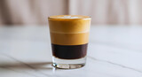 A layered coffee drink in a glass sits on a marble table