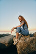 © SHOTPRIME STUDIO - smiling woman sitting on rocks by the sea wearing casual blue outfit and sneakers during sunset with clear sky and warm light