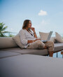 © olezzo - Attractive woman 40 years old in a white shirt sitting on a gray sofa working on a laptop on the terrace overlooking the green jungle on a bright sunny day