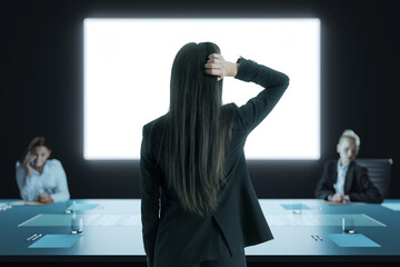 Poster - Businesswoman standing in a dark conference room thinking during meeting with back view facing bright screen and businesspeople seated at table.