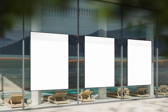 Three vertical blank poster mockups displayed on glass wall of modern building with minimalist poolside lounge chairs beneath them in natural daylight. 3D Rendering