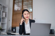 © MINAE - Young businesswoman smiling while talking on a mobile phone and working on a laptop at her modern office desk, consulting with a client
