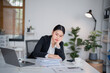 © MINAE - Asian businesswoman resting head on hand and looking down at stack of paperwork on desk while working with laptop computer in office, feeling tired, bored, and stressed from work